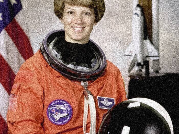 An astronaut in a flight suit beside a flag.